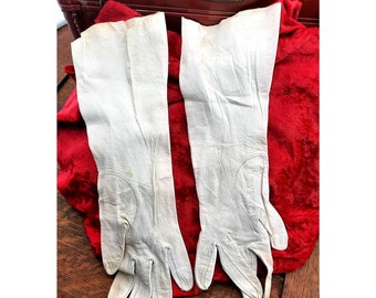 May include: A pair of white leather gloves, lying on a red velvet surface. The gloves are long and have a simple design.