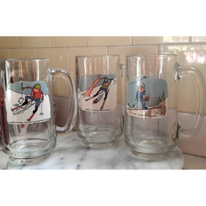 May include: Three clear glass mugs with handles. Each mug features a cartoon illustration of a person skiing. The mugs are on a white tiled surface. The text on the mugs reads: "MASHED POTATOES - Was, heavy sauce, generally hot but to slow", "ONCE - A very mountainous slid down", and "DAD - A big grin, lots of snow down".