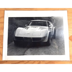 May include: A black and white photograph of a white Chevrolet Corvette sports car. The car is facing the camera and has a black number plate that reads "RACER X".