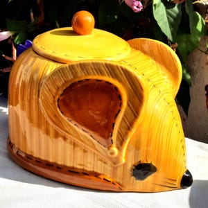 May include: A ceramic mouse-shaped cookie jar with a yellow and brown color scheme. The jar features a lid with a small orange knob. The mouse has large ears, a black nose, and a stitched design. The jar is designed to resemble a mouse.