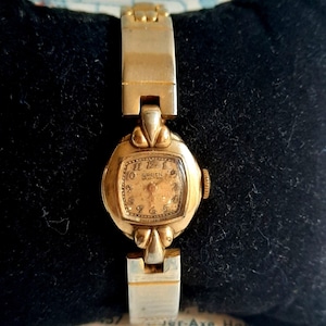 May include: A gold-toned wristwatch with a square face and a gold-toned band. The watch face has black Roman numeral hour markers and a white dial with black hands. The watch face also has the text "GRUEN VERITHIN" printed on it.