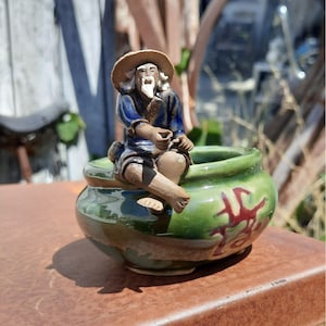 May include: A green ceramic planter with a small, hand-painted figurine of a man sitting on the rim. The figurine is wearing a blue and white robe and has a white beard. The planter has red Chinese characters on the side.