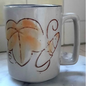 May include: White ceramic coffee mug with a brown and orange leaf design. The mug has a handle and is decorated with a brown vine.