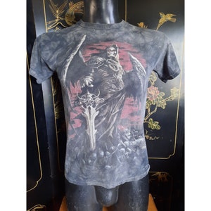 May include: A gray tie-dye t-shirt with a graphic print of a skeletal figure with wings holding a sword. The figure is standing in front of a red and black background with a crowd of smaller figures at the bottom.