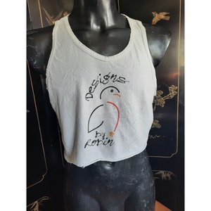 Op de afbeelding: Een wit tanktop met een gecropte zoom. Het tanktop heeft een afbeelding van een vogel met de tekst "Designs by Robin" erop gedrukt.