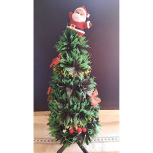 Puede incluir: Un pequeño árbol de Navidad artificial decorado con lazos rojos y oropel dorado. Una figura de Papá Noel se sienta en la cima del árbol.