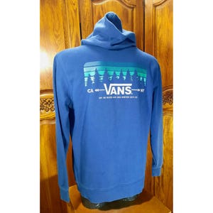 Può includere: Una felpa con cappuccio blu con una stampa grafica di una scena di foresta e il testo "VANS" in bianco. I testi "CA" e "NY" sono su entrambi i lati del logo Vans. Il testo sotto il logo recita "Off the beaten path since nineteen sixty-six".
