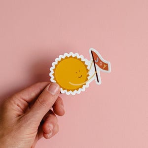 May include: A yellow sun sticker with a smiling face holding a flag that says "Joy".