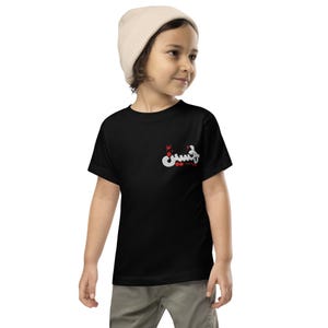 May include: A black t-shirt with white Arabic script and red accents on the upper left chest. The child in the image is wearing a beige beanie. The t-shirt is a short-sleeved style.
