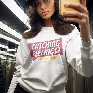 Puede incluir: Sudadera blanca con un gráfico rosa y amarillo que dice "Catching Feelings? Not my type."