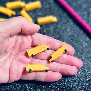 May include: A hand holding several small, yellow toy school buses with black wheels. Other buses are scattered in the background. A pink pencil is in the upper right corner. The background is a dark, textured surface.