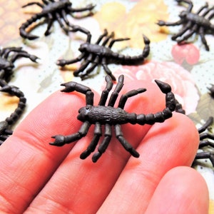 May include: A collection of small, black plastic scorpion figurines. The scorpions are detailed with segmented bodies, claws, and curved tails. They are scattered on a floral patterned surface.