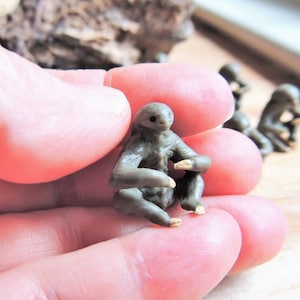 May include: A small, grey sloth figurine in a sitting pose. The figurine is made of a smooth, textured material and has a realistic appearance.