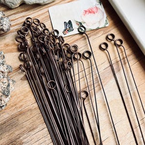 May include: A collection of long, thin, dark brown metal wire pins, each featuring a small circular loop at one end. The pins are arranged in a cluster, with several individual pins extending outward to the right side of the frame.