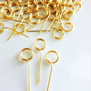 May include: A pile of gold-colored metal wire loops with a long, straight wire extending from each loop. Three of the loops are isolated and stand upright on a white surface.