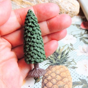 May include: A miniature plastic evergreen tree with a brown base. The tree is about 2 inches tall and is a dark green color.