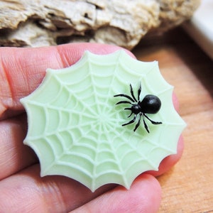 May include: A light green, web-shaped decoration with a black spider on top. The spider has eight legs and a round body. The web has a detailed, textured design. The decoration is likely made of resin or a similar material.