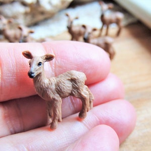 May include: A miniature brown deer figurine with detailed features, held in a hand. Other similar figurines are visible in the background. The deer has a light brown coat and small ears, perfect for a collection or display.