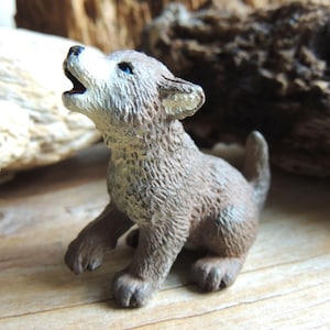 May include: A small, gray and brown wolf figurine with a white chest and blue eyes, howling with its mouth open. The figurine is sitting on a wooden surface.