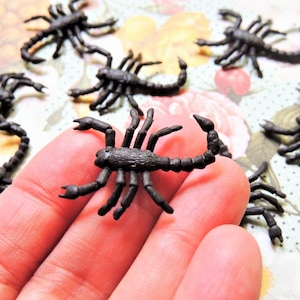 May include: A collection of eight black plastic toy scorpions. The scorpions are posed in various positions on a floral patterned surface and a hand.