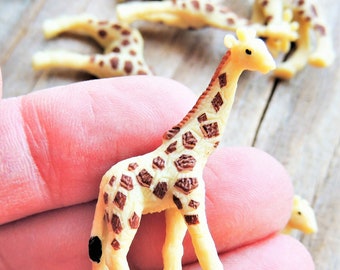 May include: A small, brown and cream colored plastic giraffe toy with a black spot on its leg. The giraffe is standing on a hand.