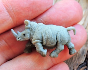 May include: A small, gray rhinoceros figurine with a detailed textured surface, a prominent horn, and small legs. The miniature rhino is held in a person's hand, showcasing its size and design. The figurine is a collectible item.