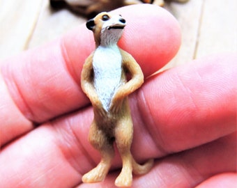 May include: A small, brown and white plastic figurine of a meerkat standing upright with its arms crossed. The figurine is about 2 inches tall.