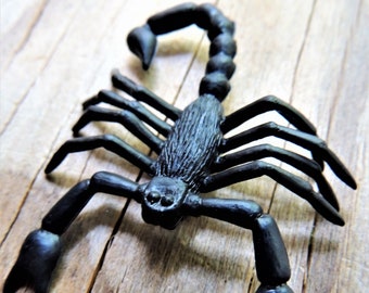 May include: A black scorpion figurine with detailed texture. The scorpion has a curved tail, multiple legs, and large pincers. The figurine is set against a wooden background.