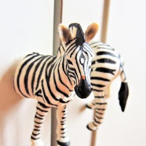May include: Two black and white zebra figurines. The zebra on the left is facing forward with its head tilted down. The zebra on the right is facing the left with its head tilted up. Both zebras are made of plastic.