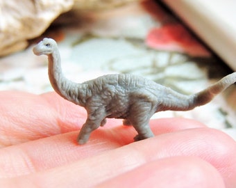 May include: A small, gray, textured dinosaur figurine with a long neck and a pointed tail. The miniature creature stands on four legs and features detailed skin patterns across its body, capturing the likeness of a sauropod-type prehistoric animal.