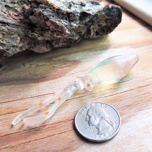 May include: A clear glass squid with orange spots and a blue stripe. The squid is about the size of a quarter.