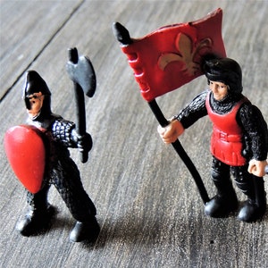 May include: Three toy knights, one holding a red flag with a fleur-de-lis, another holding a sword, and the third holding a shield with a red heart.