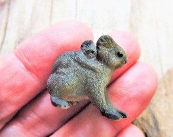 May include: A small, detailed figurine of a koala with a baby koala on its back. The figurine is primarily olive green with black accents on the paws and ears. The koala is in a sitting position.