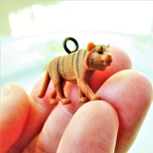 May include: A small, brown and black tiger figurine with a metal loop on its back. The figurine is being held in a person's hand.