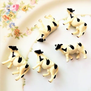 May include: Five black and white plastic toy cows on a white plate with a floral design. The cows are in various poses.