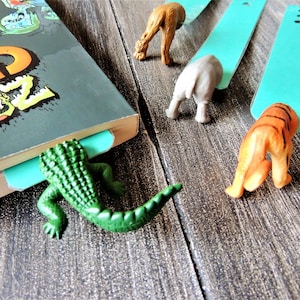 May include: A book with a green crocodile bookmark, a blue bookmark with a gray elephant, a blue bookmark with a brown camel, and a blue bookmark with an orange tiger. The bookmarks are all on a wooden surface.