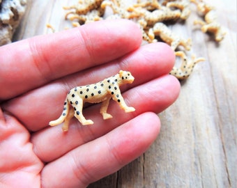 May include: A small plastic cheetah figurine with black spots on a tan body. The figurine is being held in a person's hand.