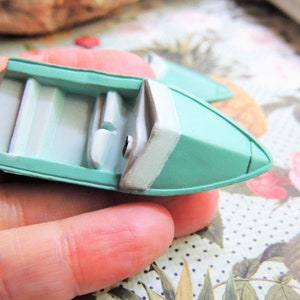 May include: A miniature, light teal boat with white interior and accents. The boat has a small, detailed design, with a white stripe along the side. The boat is a decorative item, possibly for crafts or display.