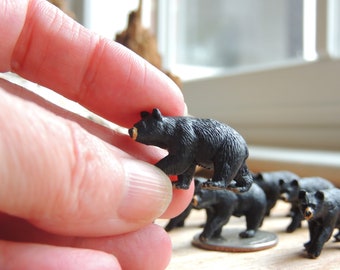 May include: A collection of miniature black bear figurines with tan snouts. One bear is held between fingers, while others stand on a surface. The bears are detailed with textured fur, suggesting a wildlife or nature theme.