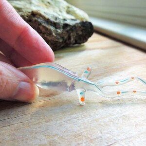 May include: A clear, translucent, sea creature figurine with blue and orange accents. The figurine has a streamlined body and long, curved appendages. The design is held by a hand, suggesting a small size and delicate nature.