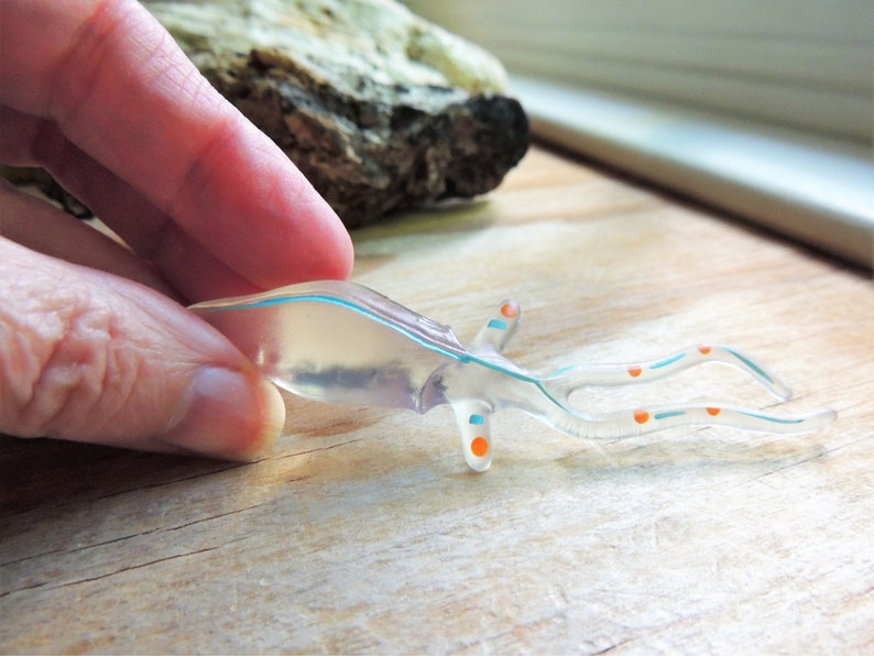 May include: A clear, translucent, sea creature figurine with blue and orange accents. The figurine has a streamlined body and long, curved appendages. The design is held by a hand, suggesting a small size and delicate nature.