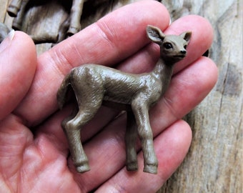 May include: A small, brown plastic deer figurine. The deer is standing with its legs extended and its head up. The figurine is held in a hand, with other similar figurines visible in the background. The deer is approximately 2 inches tall.