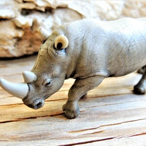 May include: A gray plastic rhinoceros toy with a white horn and small ears. The toy is posed in a walking position on a wooden surface. The rhino's skin has a textured appearance.
