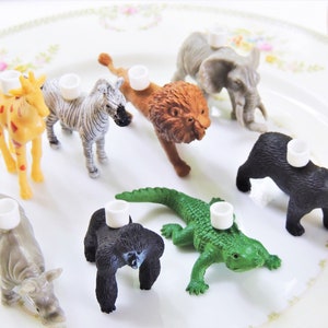 May include: A set of eight animal figurines with white candle holders on their backs. The animals include a giraffe, zebra, lion, elephant, gorilla, crocodile, rhino, and bear. The figurines are made of plastic and are painted in realistic colors.