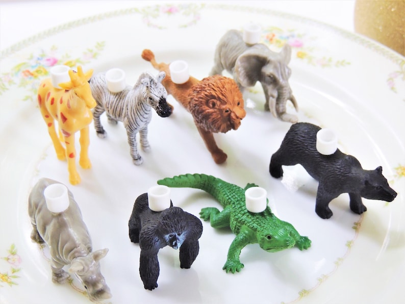 May include: A set of eight animal figurines with white candle holders on their backs. The animals include a giraffe, zebra, lion, elephant, gorilla, crocodile, rhino, and bear. The figurines are made of plastic and are painted in realistic colors.