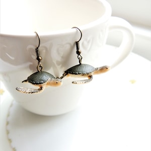May include: A pair of bronze turtle earrings with a green shell and a beige body. The earrings are hanging from a white cup.