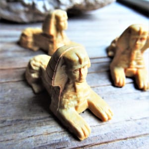 May include: Three small, beige, ceramic sphinx figurines. The sphinx figurines are in a seated position with their paws outstretched. The figurines are on a wooden surface.