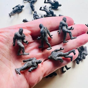 May include: A collection of small, dark gray plastic figurines depicting Bigfoot or Sasquatch in various walking and standing poses. The figures feature textured surfaces to represent fur and are scattered across a flat, light-colored surface.