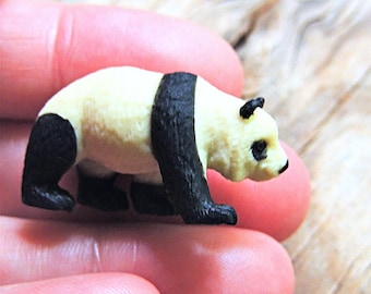May include: A small, black and white panda figurine. The panda is standing on all fours and has a black nose and eyes.