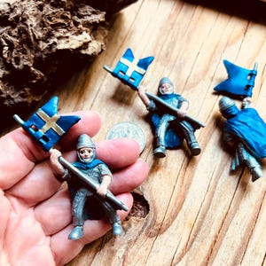 May include: Miniature toy soldiers in silver armor and blue capes, holding blue flags with gold crosses. The figures are on a wooden surface, with a coin visible. The flags are a vibrant blue, and the soldiers have detailed features.